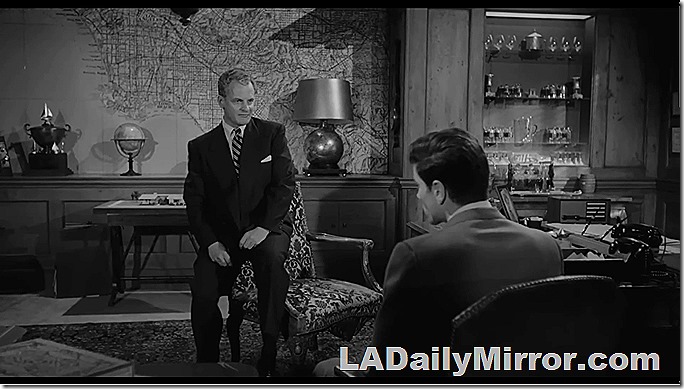 An office. Man in a suit is sitting on the arm of a chair. A huge map of Los Angeles is on the wall. The office also has an elaborate bar. Also Back of the Head Guy